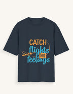 Front view of a dark navy blue unisex oversized streetwear t-shirt featuring a stylized airplane graphic and the text "CATCH FLIGHTS NOT FEELINGS" in orange and teal lettering across the center.